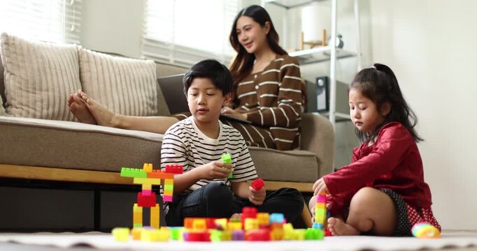Asian mother working on a laptop from home, while her young son and daughter drawing at a coffee table while raising hands, signifying active child education and creative homeschooling