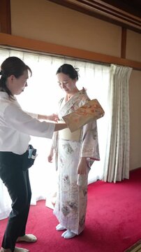 Woman smiles in traditional kimono, standing beside a red carpet. Helper adjusts her garment, ensuring perfect fit and style. Soft light from window highlights delicate floral patterns