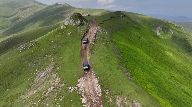 Drone shot of off-road buggies driving along a dirt path through picturesque green mountain hills. Cinematic landscape featuring rolling alpine meadows and scenic mountain ranges