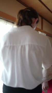 Woman in a kimono receives help from another. The helper adjusts her garment with care. Soft light bathes the traditional room. Both appear calm and focused. Ideal for cultural, bridal
