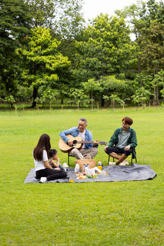 GrandFather playing acoustic guitar for child during picnic. Asian family enjoy outdoor picnic with food and drinks in park sunny day, outdoor activitie with daughter father, mother grandfather