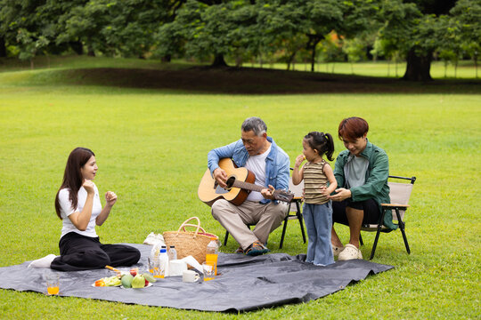 GrandFather playing acoustic guitar for child during picnic. Asian family enjoy outdoor picnic with food and drinks in park sunny day, outdoor activitie with daughter father, mother grandfather