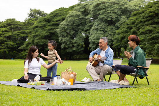 GrandFather playing acoustic guitar for child during picnic. Asian family enjoy outdoor picnic with food and drinks in park sunny day, outdoor activitie with daughter father, mother grandfather