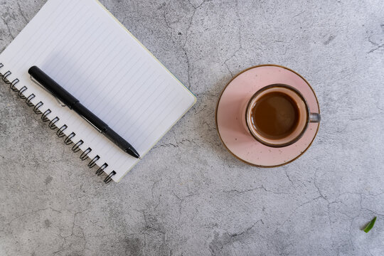 White notebook rests beside a pink saucer. Black pen lies beside lined pages. Coffee cup holds warm, dark brew. Gray surface shows subtle texture. Stillness suggests quiet work or thought