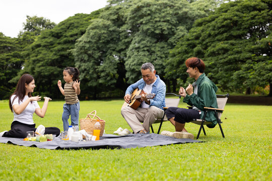 GrandFather playing acoustic guitar for child during picnic. Asian family enjoy outdoor picnic with food and drinks in park sunny day, outdoor activitie with daughter father, mother grandfather