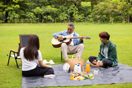 GrandFather playing acoustic guitar for child during picnic. Asian family enjoy outdoor picnic with food and drinks in park sunny day, outdoor activitie with daughter father, mother grandfather