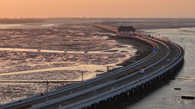 Aerial view of a curving coastal causeway bridge at sunset stretching across mudflats and tidal pools with light reflecting on the roadway and sparse traffic through an expansive low tide landscape