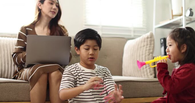 Asian mother working on a laptop from home, while her young son and daughter drawing at a coffee table while raising hands, signifying active child education and creative homeschooling