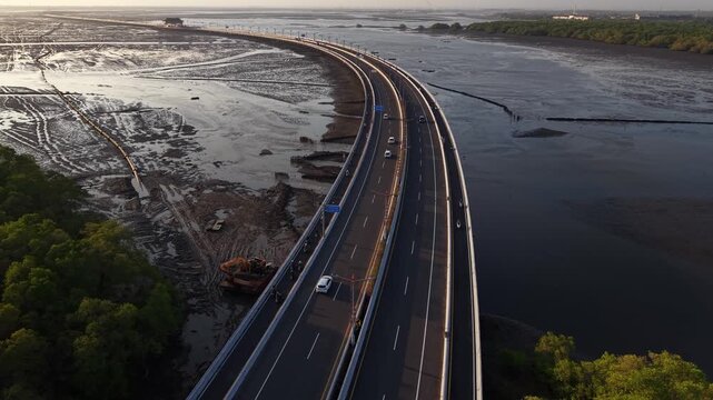 Aerial shot of a curving coastal highway and toll bridge crossing tidal mudflats with sparse traffic, mangrove shoreline and dramatic evening light, illustrating coastal infrastructure and transport