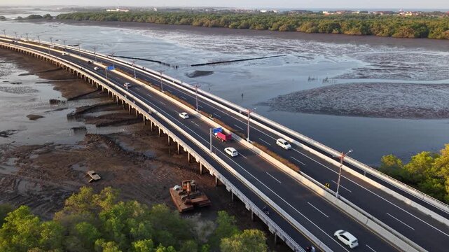 Aerial shot of a modern elevated highway bridge carrying light traffic over tidal mudflats and mangrove shoreline at low tide, showing coastal infrastructure, natural wetlands and calm late afternoon