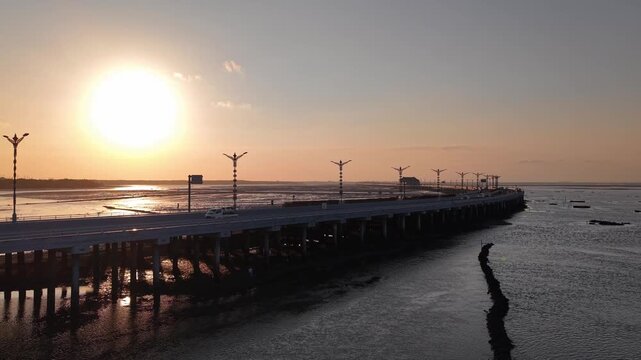 Golden sunset lights a long coastal causeway bridge, casting strong reflections on muddy tidal flats and creating silhouetted lamp posts and cars against a calm sea and expansive horizon