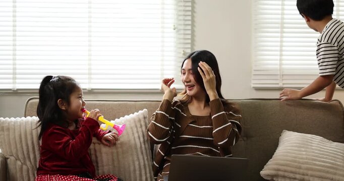 Asian mother working on a laptop from home, while her young son and daughter drawing at a coffee table while raising hands, signifying active child education and creative homeschooling