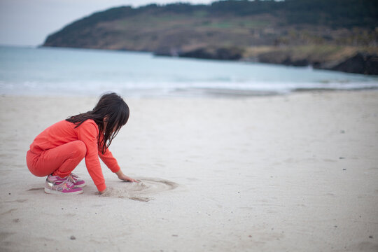 Side view of a little girl drawing in the sand on a beautiful white sand beach with sea background, her face covered by hair.