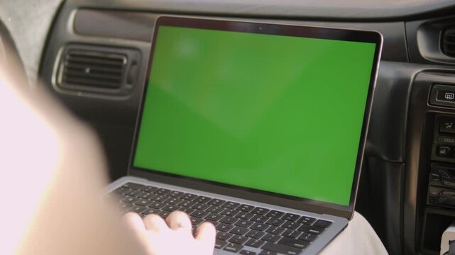 Person uses laptop with green screen inside car. Hands type on keyboard, focused on screen. View shows dashboard and car interior. Good for travel, remote work, or digital content overlays