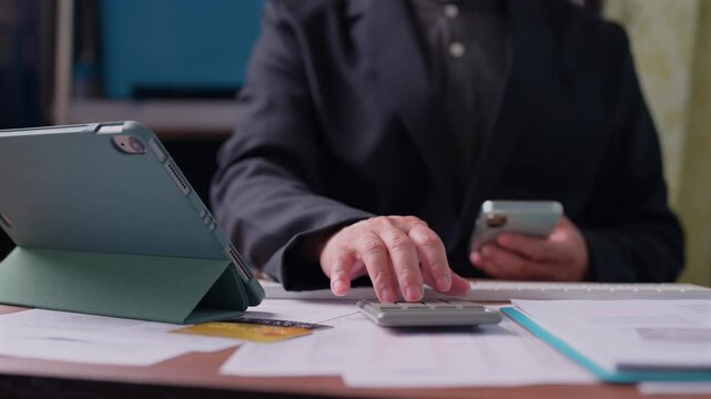 Professional accountant calculates financial data, presses calculator keys, holds mobile phone, sits beside tablet, credit card, documents, analyzing budget expenses inside office workspace.