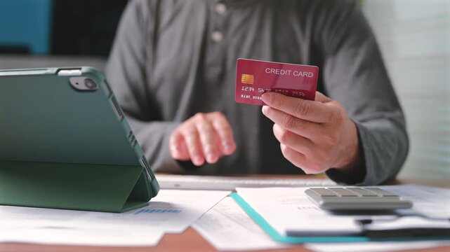 Man holding his red credit card types computer keyboard inside home office workspace making online payment transaction near tablet calculator paper document managing personal finance.