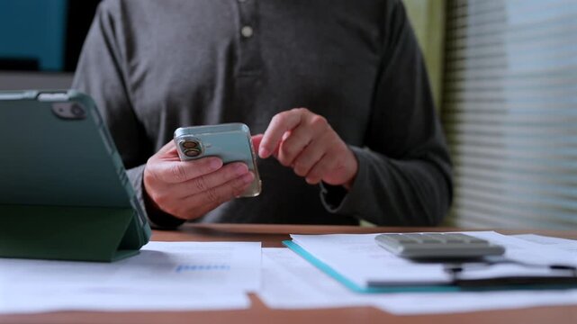 Businessman holding smartphone reviews financial paperwork near digital tablet calculator inside workspace, managing budget, tracking expenses, calculating investment revenue, analyzing data.