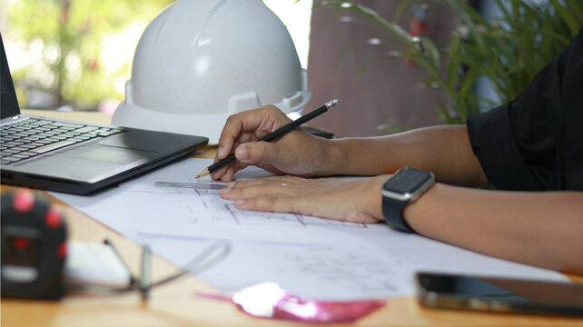 Close-up of an architect drafting residential blueprints with ruler and pencil beside a safety helmet and laptop. Engineering, construction planning, architectural , and building development concept.