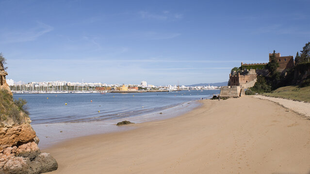 The historic Fort of S&atilde;o Jo&atilde;o do Arade stands beside the wide, sandy shores of Praia da Angrinha in Ferragudo, Algarve under a clear blue sky.