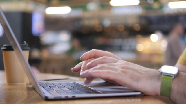 Hands type on laptop in cozy cafe. Warm light glows softly around. Coffee cup rests beside keyboard. Person wears smartwatch and yellow shirt. Scene captures relaxed work or study moment