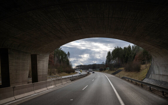 A view from beneath a massive concrete highway bridge framing a road stretching through a landscape with trees and distant vehicles under an overcast sky