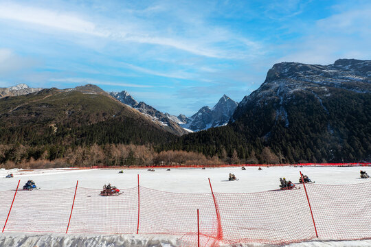 People enjoying snowmobiling on a wide snow-covered field surrounded by majestic snow-capped mountain peaks and evergreen forests under a bright blue sky with light clouds