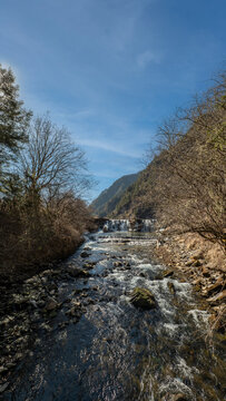 scenic mountain stream with rushing water, rocky rapids, and bare trees along the banks leading to a small waterfall under a clear blue sky on a bright day
