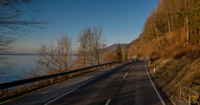 A scenic coastal road stretching alongside a calm lake with bare trees and a forested hill under a clear blue sky on a sunny day during a road trip