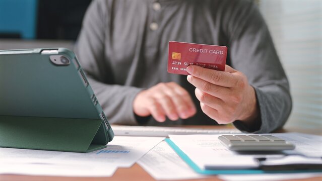 Man holds red credit card, types tablet keyboard, calculates business expenses, manages personal finance, pays bills online, performs electronic commerce transactions inside home workspace office.