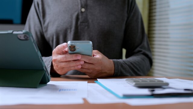 Man holds mobile phone, reviewing financial document, calculator, tablet computer over wooden desk inside workspace, managing personal budget, tracking investment portfolio, calculating expenses.