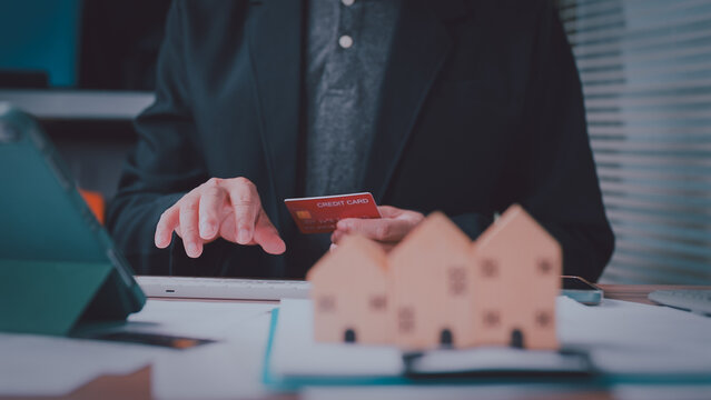 Businessman holding credit card operates keyboard near wooden houses, processing online mortgage payment, buying property, managing real estate finance, conducting digital banking transaction.