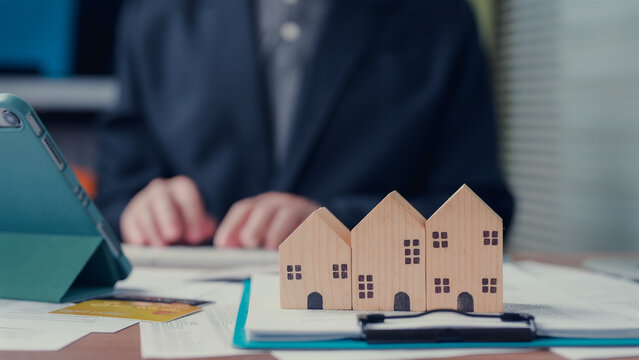 Agent types keyboard beside wooden house models, reviewing financial documents, credit card, tablet inside office workspace, planning mortgage investment loan budget cost rates.