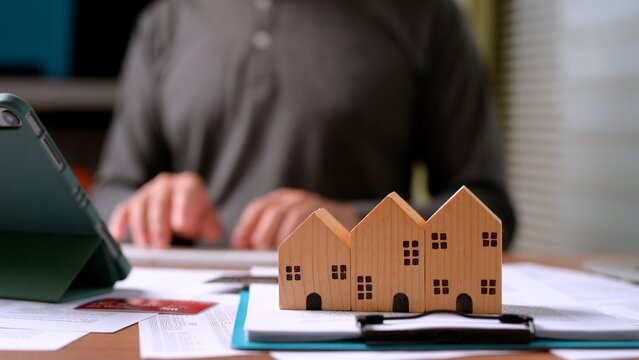 Small wooden house models rest atop financial paper documents businessman works background, calculating mortgage payment, real estate investment, property tax planning, home insurance cost.