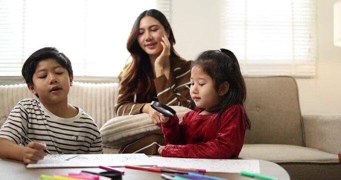 Asian mother remote working from home on a laptop in the living room while her two children are drawing and coloring at a low table, balancing work and family life