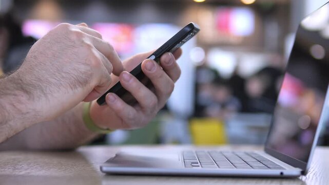 Hand uses smartphone while laptop sits open nearby. Person appears focused and engaged with device. Suitable for tech, work, or multitasking themes. Blurred background suggests public or cafe setting