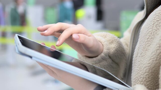 Person uses tablet at airport. Fingers tap screen. Background shows blurred travelers. Yellow tape visible. Light-colored coat worn