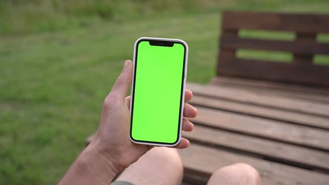 Hand holds smartphone with green screen. Person sits on wooden bench outdoors. Background shows grass and nature. Ideal for app or advertisement mockups