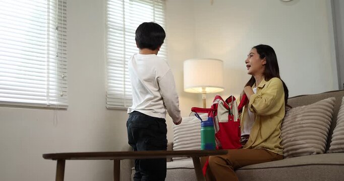 Asian mother and son preparing for school in a bright home setting, with the parent helping the child pack his red backpack and water bottle, showcasing family support and education responsibilities