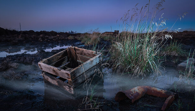 An aged wooden crate rests in a muddy swamp surrounded by tall wild grasses under the dim twilight sky as a discarded metallic pipe lies partially submerged within the murky marshland