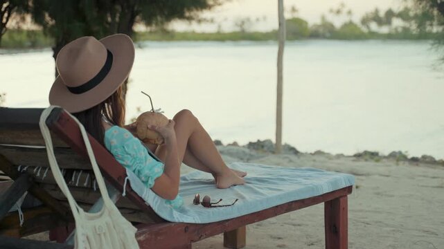 Woman relaxing on a wooden beach lounge chair while holding a coconut drink and wearing a fedora hat by the calm waters during her serene tropical vacation under the clear and bright summer sky.