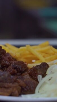 Slow motion camera panning of traditional roasted beef meat served with crispy french fries and sliced raw onions on a white plate, vertical video.