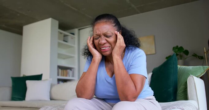 African American senior woman holding temples, wincing from pain, lifting head, looking up on couch