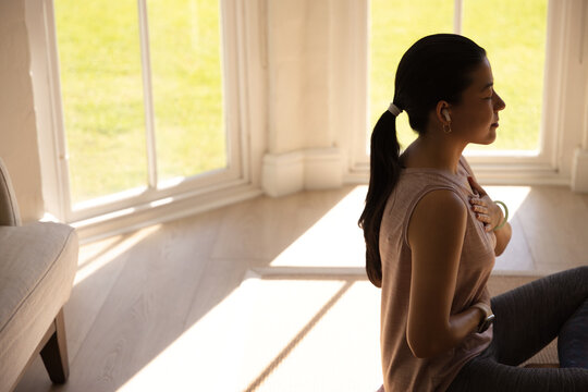 Asian woman meditating by bay window hands on abdomen, wearing tank and leggings, watch, copy space