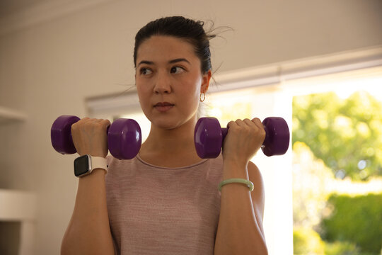Asian woman lifting purple dumbbells at shoulders by window, wearing sleeveless top and smartwatch