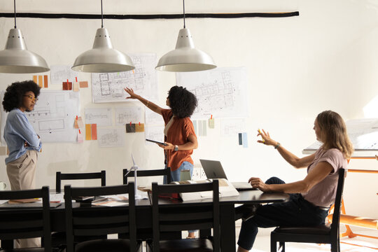 Diverse female colleagues reviewing layout plans in bright design studio with laptop and tablet