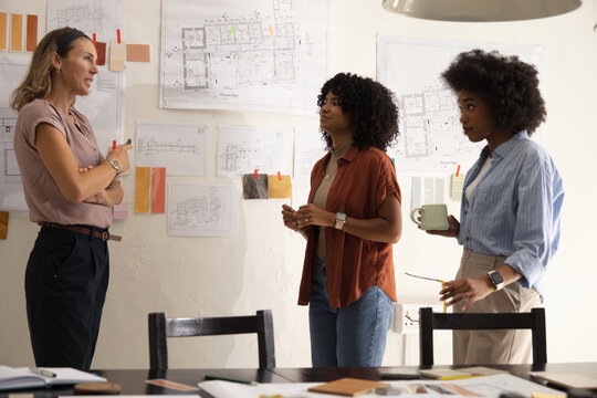 Diverse female colleagues discussing pinned plans and color swatches at studio with paper samples