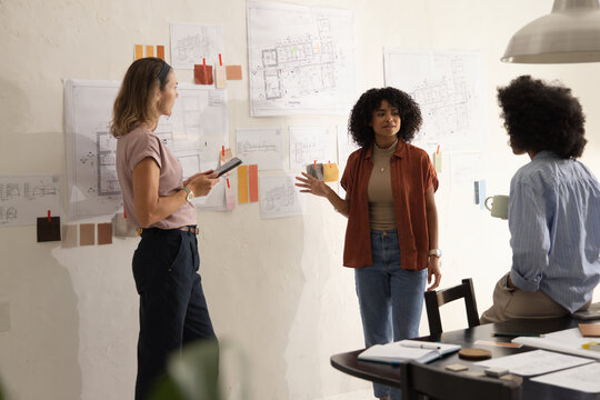 Diverse female colleagues discussing floor plans, gesturing toward swatches, using tablet in studio