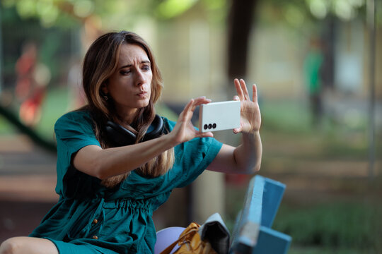 Surprised Woman Sitting On Park Bench Recording Unexpected Scene On Phone. Astounded  witness focused on filming hidden outdoor event with smartphone while resting on bench