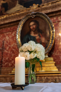 Lit white pillar candle and a vase of white hydrangeas on an altar cloth in front of a gilded oval painting of Christ.
