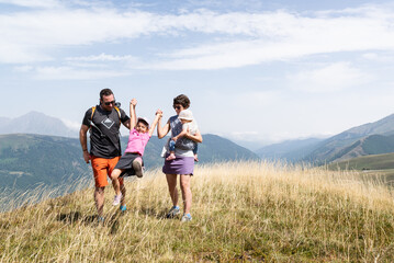 Fototapeta premium Cheerful mother with her baby in a carrier and father holding their young daughter's hands, swinging and lifting her in the air on a mountain trail in Peyragudes, French Pyrenees.